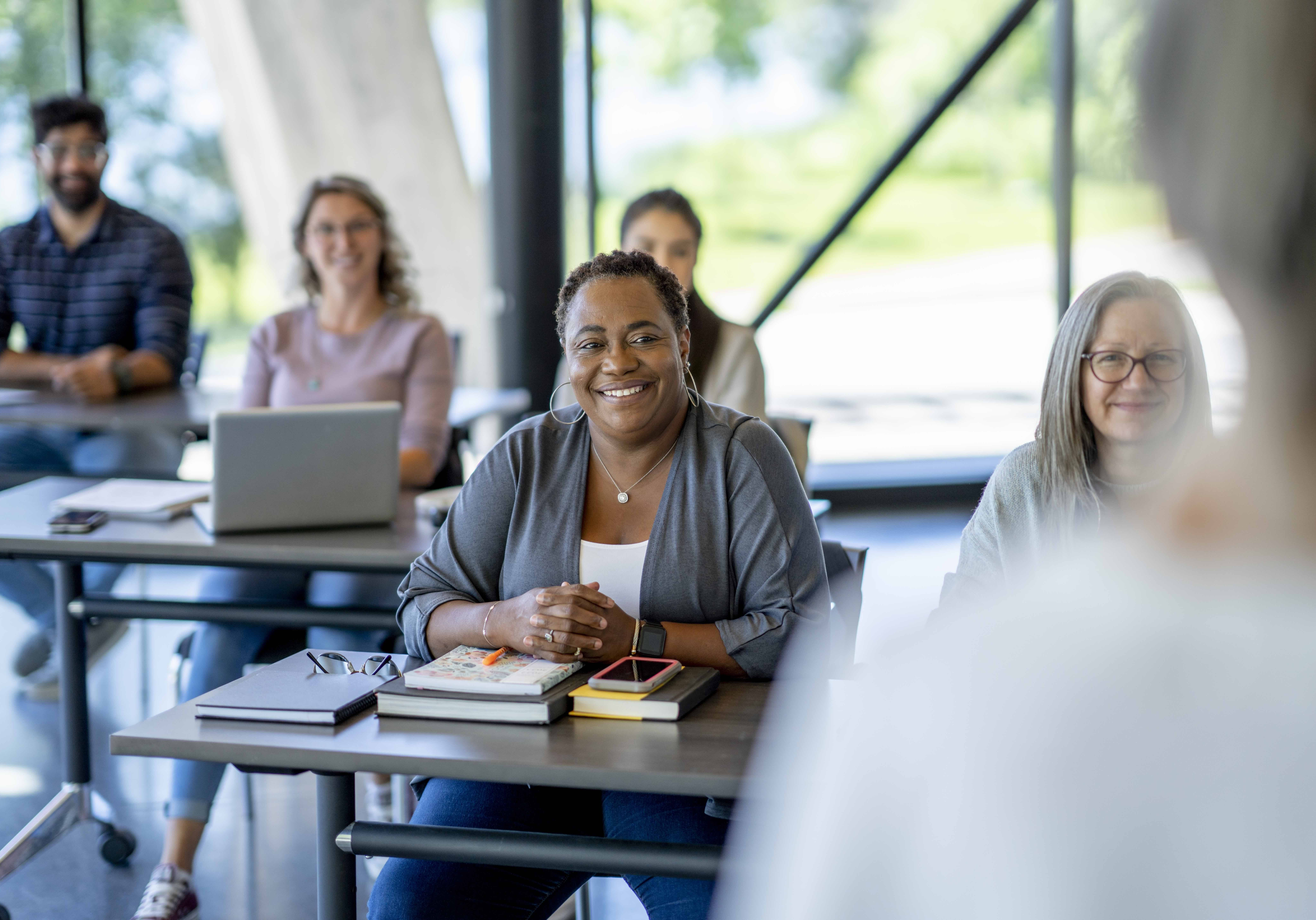 A class of mature students sit in class together as they listen attentively to the teacher.  They are each dressed casually and have laptops and books out in front of them as they take notes during the teaching.