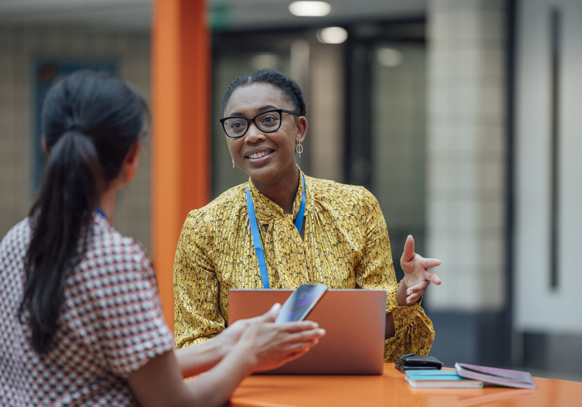 Two female teachers sitting at a table, having a discussion in the lobby area in the school they work at in Gateshead, North East England. They are talking about school issues together while using a laptop.